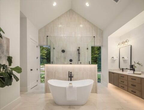Luxurious white bathroom with a stand-alone bathtub and glass shower with large windows behind it showing the trees in the background.